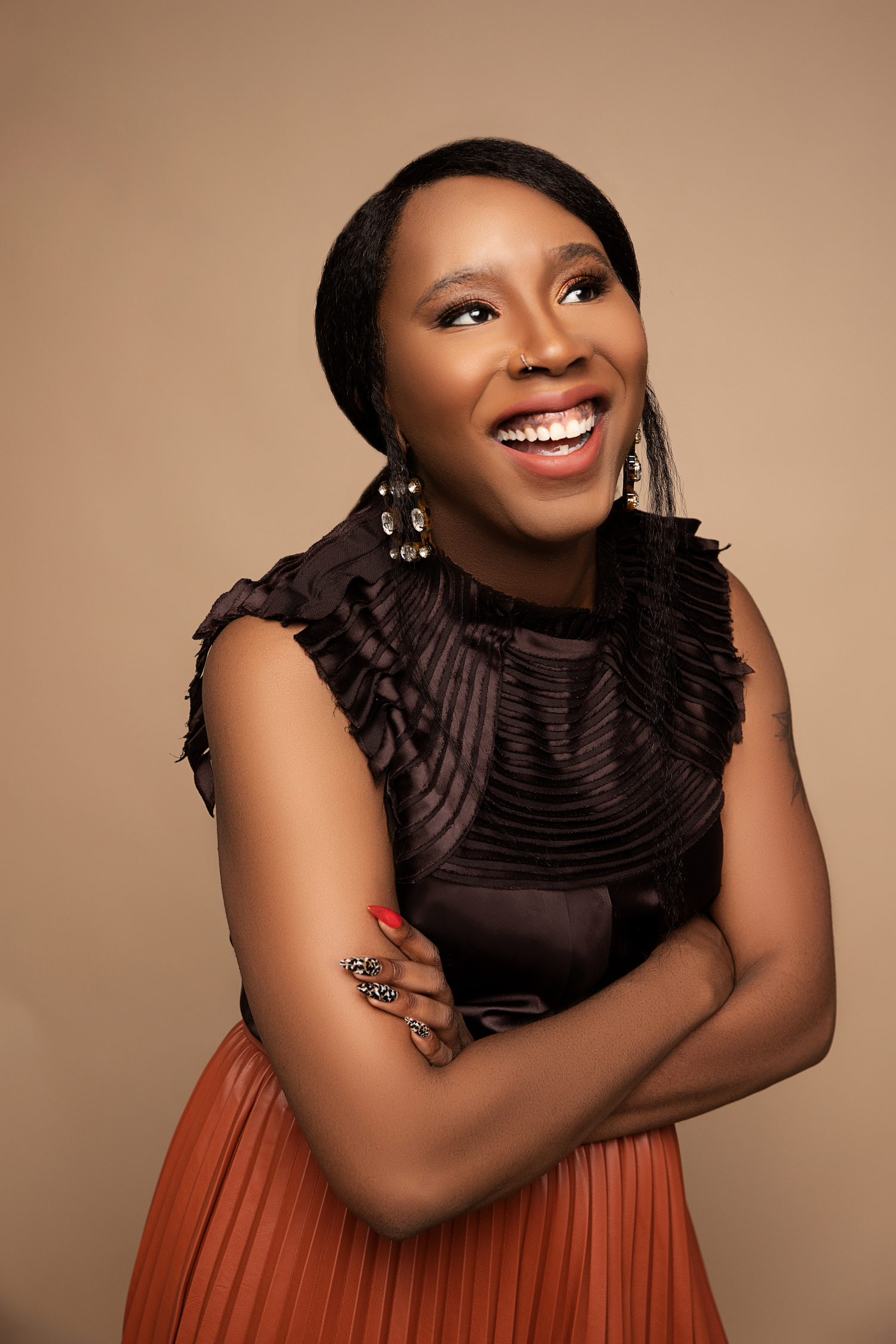 Smiling Black woman in brown ruffled top and orange pleated skirt, arms crossed, beige background